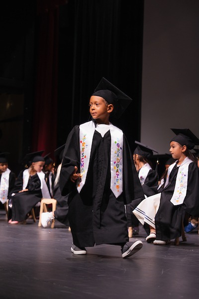 A young child in a graduation cap and gown walks on stage, wearing a sash that reads "Preschool Graduate," while other children in similar attire sit nearby.