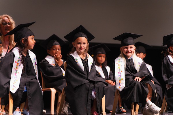 Young children in graduation gowns and caps sit on stage, smiling and looking toward the audience, celebrating their preschool graduation.