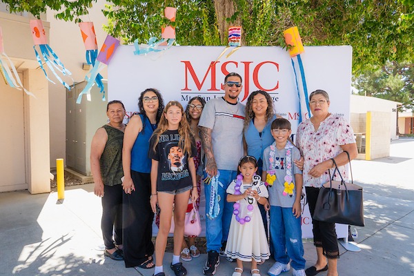 A group of people, including children and adults, pose together outdoors in front of a banner for MSJC, with colorful paper decorations hanging above them.
