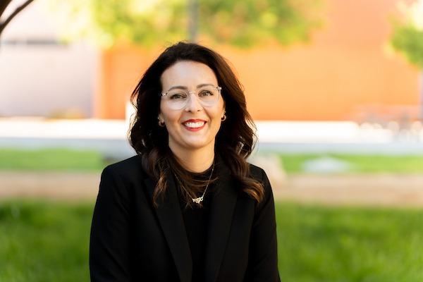 A smiling person with dark hair and glasses wears a black blazer and necklace, standing outdoors in front of green grass and a blurred orange wall.